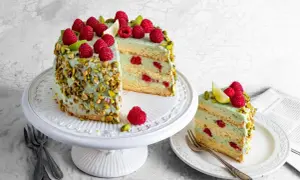Wyjątkowy tort pistacjowo malinowy, udekorowany świeżymi malinami i pistacjami. Kawałek na talerzyku kusi warstwami kremu i owoców.