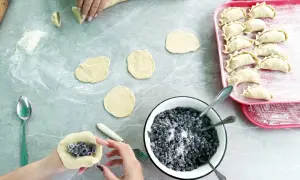Ręce formują ciasto wonton z jagodami i cukrem. Na blacie leżą okrągłe placki ciasta, miska z jagodami i tacka z gotowymi pierożkami.