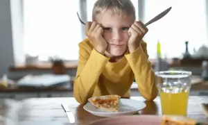 Chłopiec z widoczną wybiórczością pokarmową trzyma widelec i nóż, patrząc z niechęcią na pizzę.