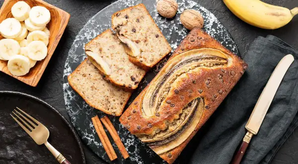 Szybkie fit ciasto bananowe z kawałkami czekolady i bananem na wierzchu, obok plasterki banana, orzechy i cynamon.