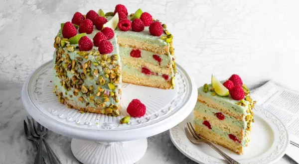 Wyjątkowy tort pistacjowo malinowy, udekorowany świeżymi malinami i pistacjami. Kawałek na talerzyku kusi warstwami kremu i owoców.