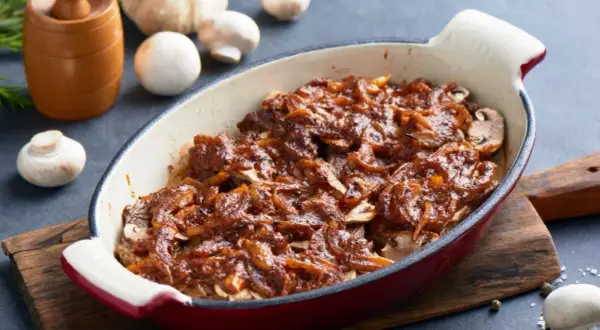 Karkówka pieczona w plastrach z cebulą i ketchupem, podana w naczyniu żaroodpornym. Obok pieczarki i czosnek.