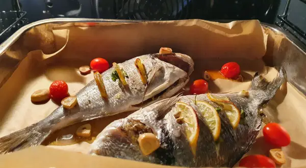 Dwie dorady ryby pieczone z cytryną, czosnkiem i pomidorkami koktajlowymi na papierze do pieczenia.