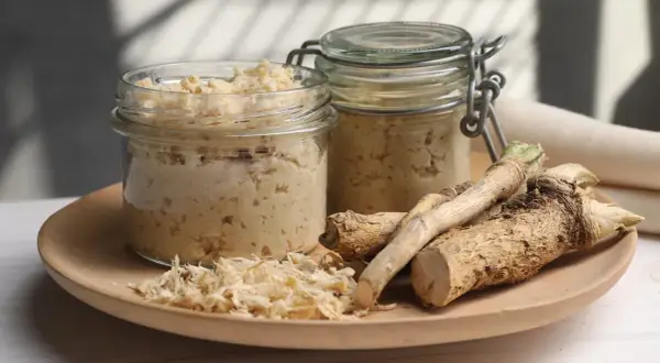 Jak zrobić chrzan domowy do słoików? Na talerzu świeży chrzan, tarta masa i dwa słoiki gotowego produktu.