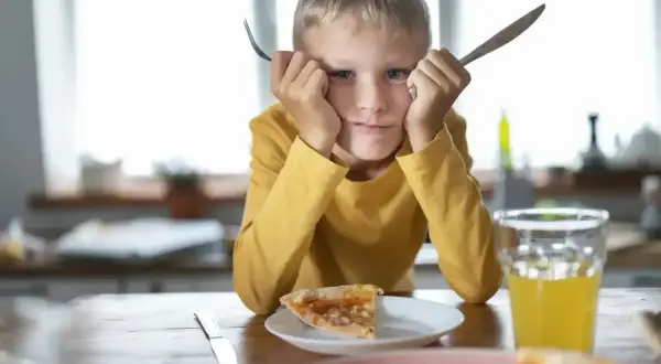 Chłopiec z widoczną wybiórczością pokarmową trzyma widelec i nóż, patrząc z niechęcią na pizzę.