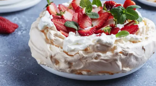 Pyszny tort bezowy z mascarpone i bitą śmietaną, ozdobiony świeżymi truskawkami i miętą.