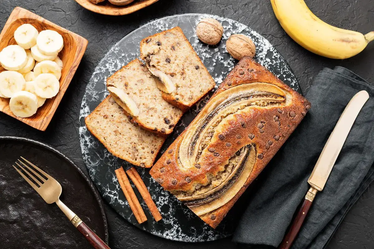 Szybkie fit ciasto bananowe z kawałkami czekolady i bananem na wierzchu, obok plasterki banana, orzechy i cynamon.