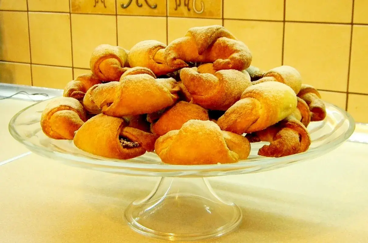Rogaliki drożdżowe z przepisu babci, posypane cukrem pudrem, piętrzą się na szklanej paterze.