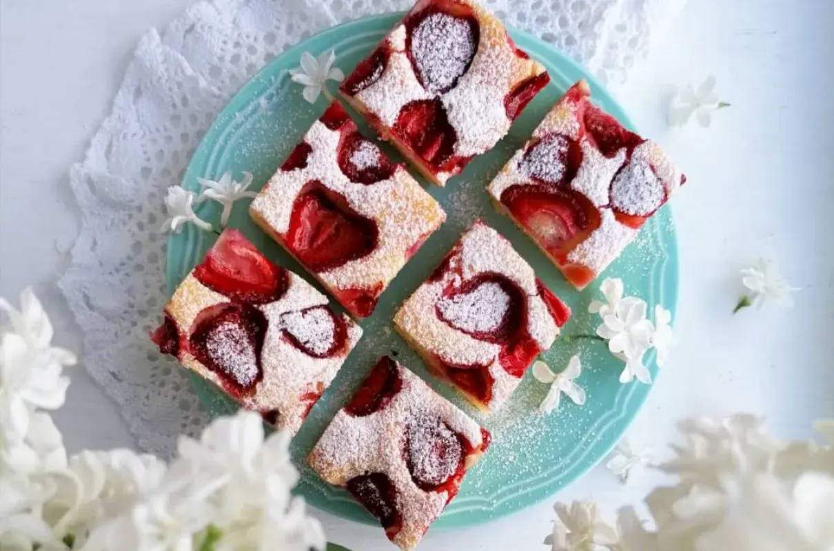 Sześć kawałków pysznego, szybkie ciasto bezglutenowe z truskawkami, posypane cukrem pudrem, na turkusowym talerzu, ozdobione białymi kwiatami.