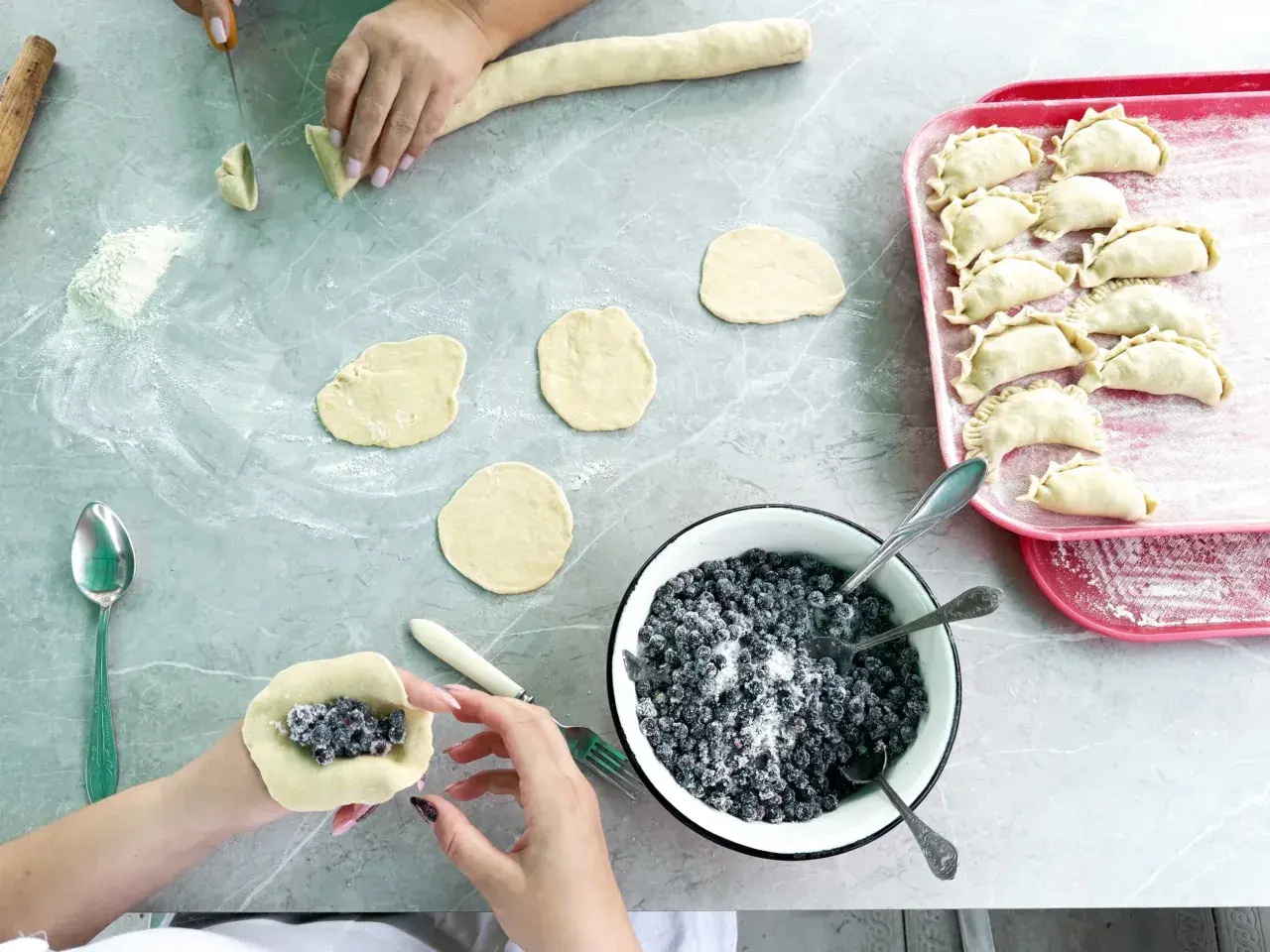 Ręce formują ciasto wonton z jagodami i cukrem. Na blacie leżą okrągłe placki ciasta, miska z jagodami i tacka z gotowymi pierożkami.