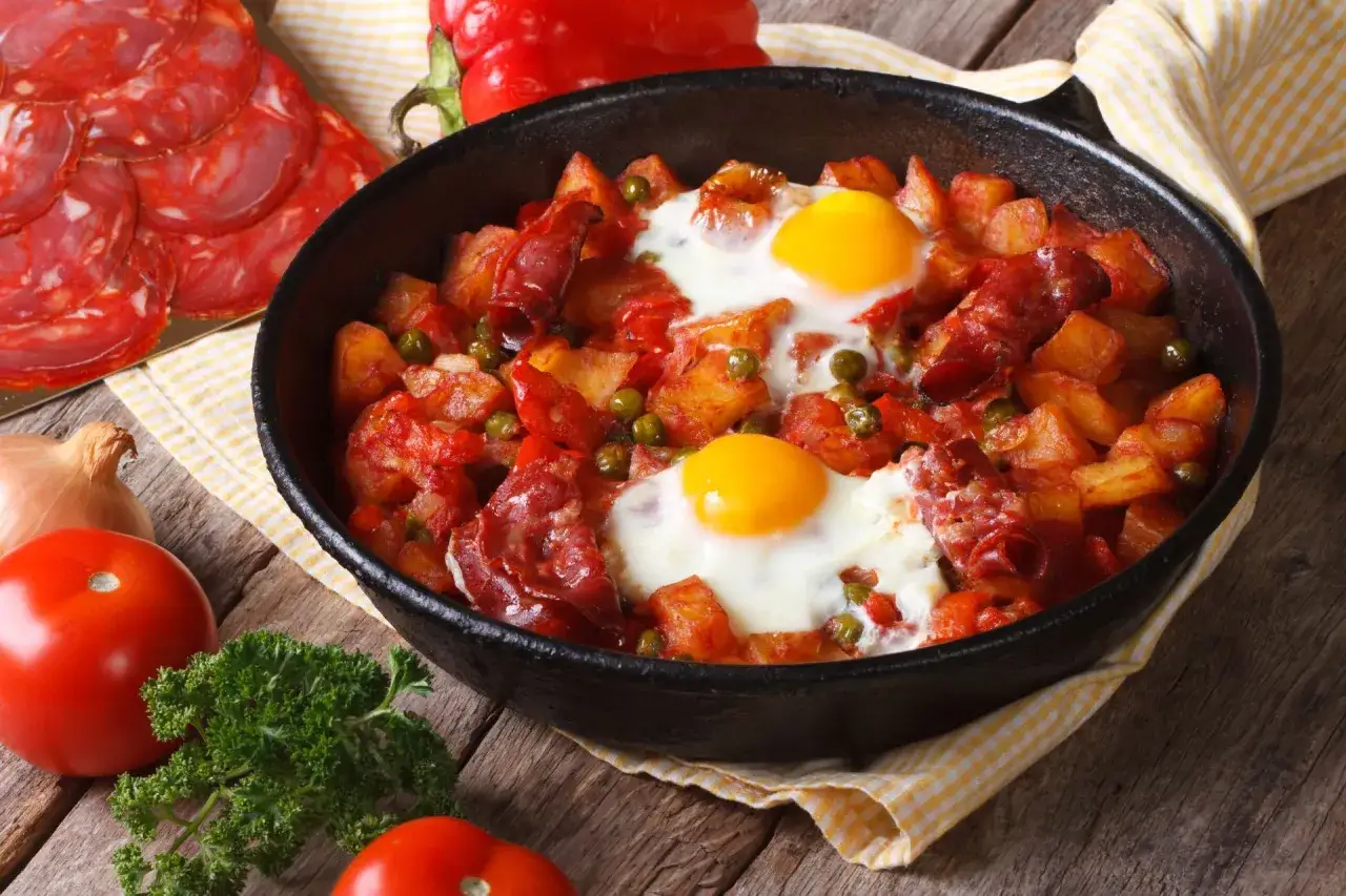 Pyszny obiad z jajek z ziemniakami, chorizo i groszkiem w patelni. Obok pomidory, cebula i natka pietruszki.