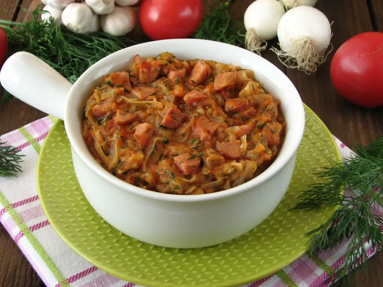 Jak ugotować dobry bigos z młodej kapusty? Aromatyczny bigos w białej misce, z dodatkiem kiełbasy i koperku. Na stole czosnek, pomidory i cebula.