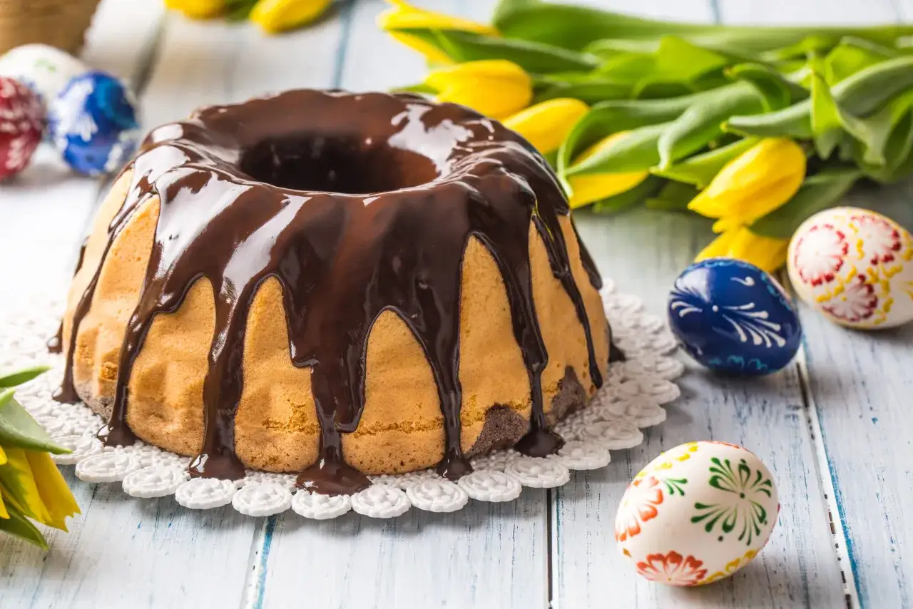 Babka z majonezem sprawdzony przepis, polana czekoladą, obok pisanki i żółte tulipany.