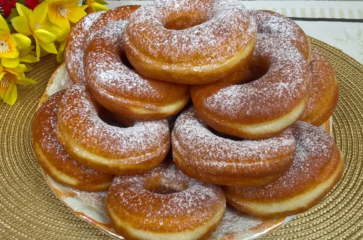 Stos puszystych oponków z cukrem pudrem, idealnych na przepis na oponki zwykłe. Obok żółte kwiaty.