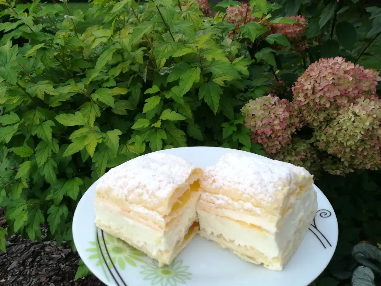 Dwa kawałki ciasta z tradycyjnym kremem do napoleonki, posypane cukrem pudrem, na talerzu na tle zieleni i kwitnących hortensji.