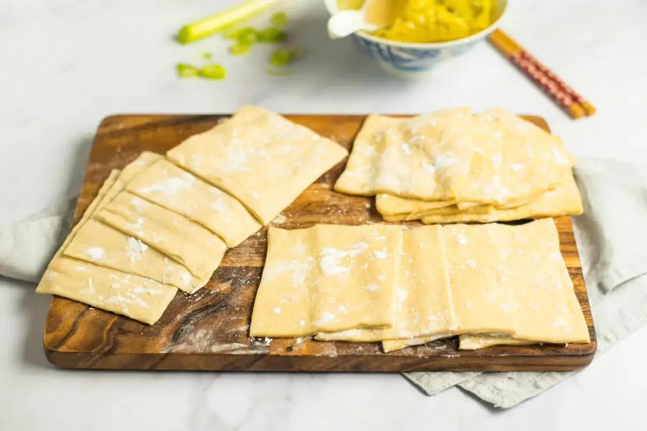 Gotowe do lepienia pierożki wonton leżą na drewnianej desce. Obok miseczka z farszem i pałeczki.
