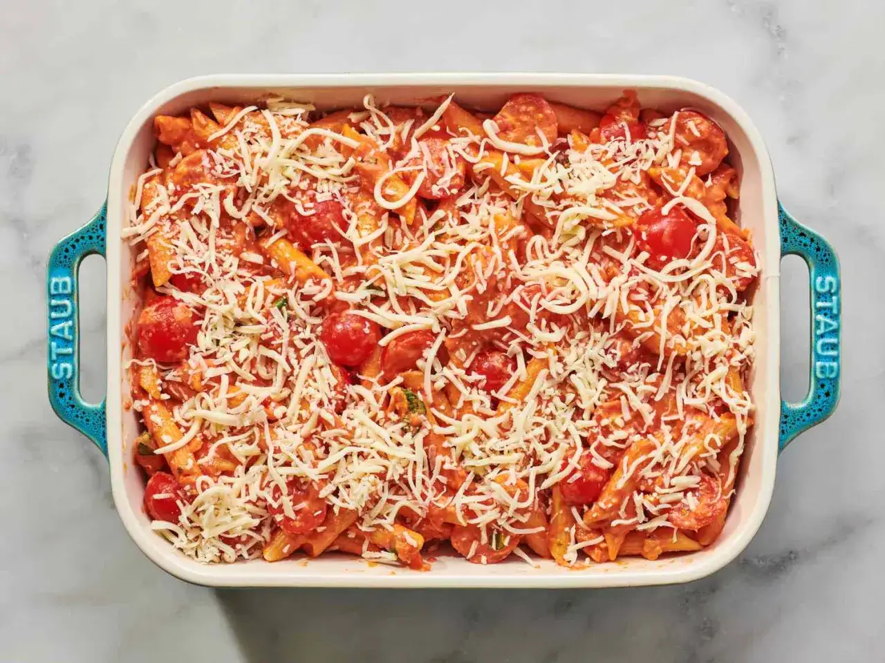 Penne z sosem pomidorowym, pomidorkami koktajlowymi i obficie posypane startą mozzarellą, gotowe do zapieczenia w naczyniu Staub.