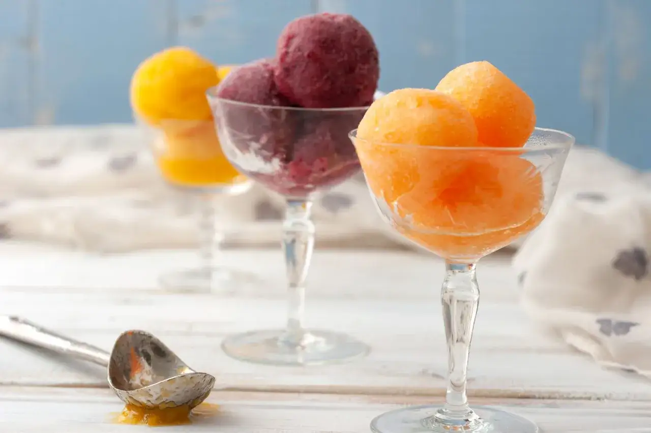 Jak zrobić sorbet? Trzy puchary z owocowymi sorbetami: pomarańczowym, jagodowym i żółtym. Obok łyżka z resztkami sorbetu.