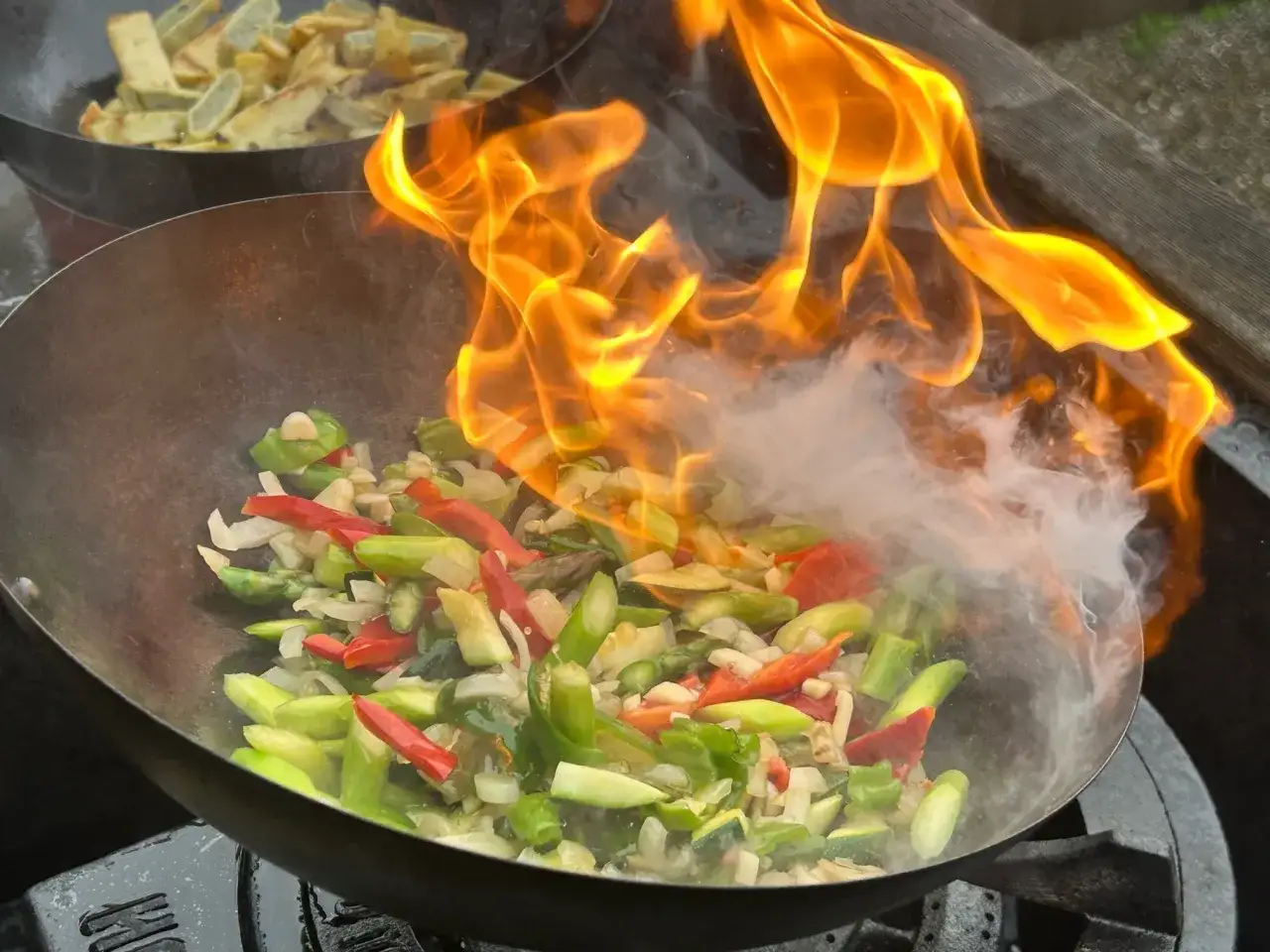 Ogień buchający z woka, w którym smaży się kolorowy stir fry z warzywami. W tle widać drugą patelnię z ziemniakami.