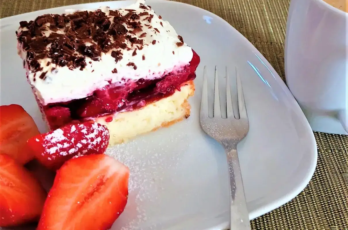 Puszysty biszkopt z truskawkami i mascarpone, posypany wiórkami czekolady. Obok świeże truskawki i widelec.