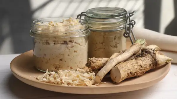 Jak zrobić chrzan domowy do słoików? Na talerzu świeży chrzan, tarta masa i dwa słoiki gotowego produktu.